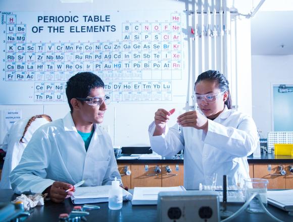 Two students working in UCSB chem lab with big periodic table on the wall behind